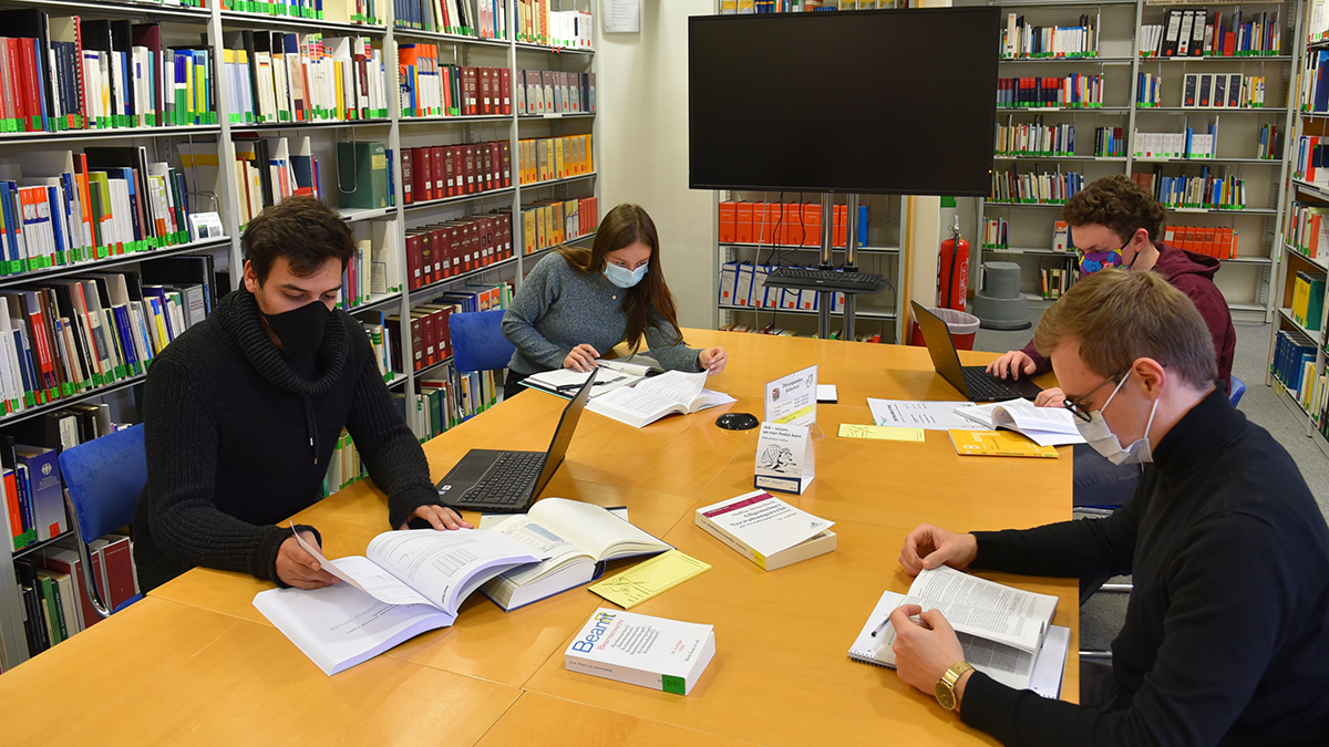 Vier junge Menschen sitzen mit Abstand und Masken an einem großen Tisch in einer Bibliothek und arbeiten mit Büchern.