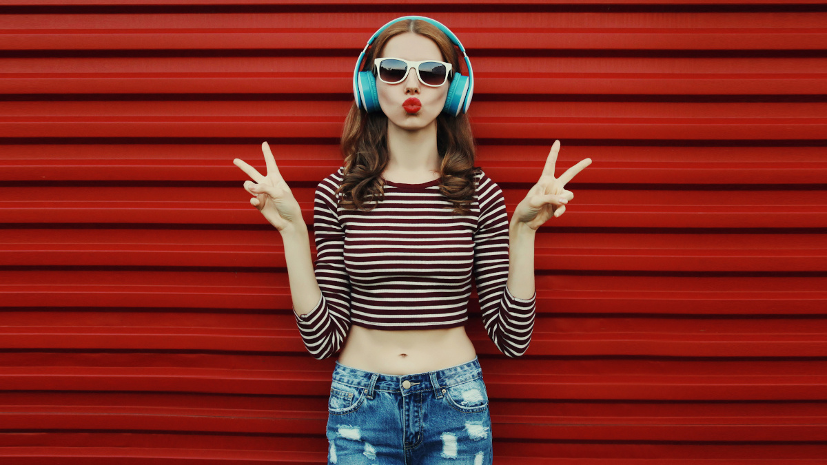 Eine junge Frau steht in in blauer Jeans und rot-weiß gestreiftem Crop-Top vor einer roten Wand. Sie trägt hellblaue Kopfhörer, eine Sonnenbrille, macht mit ihrem Mund ein Duckface und zeigt mit ihren Händen jeweils ein Peace-Zeichen.
