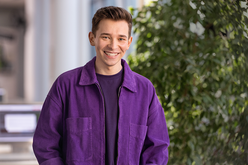 Portraitfoto von Kevin: Ein junger Mann mit braunen Haaren, grauen Augen lächelt und trägt eine lila Jacke über einem lila Shirt.